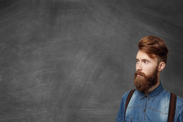 Ritratto di studentessa bruna alla moda con barba sfocata che indossa camicia di jeans e bretelle che guarda lontano in lontananza avendo un'espressione seria e fiduciosa sul viso.