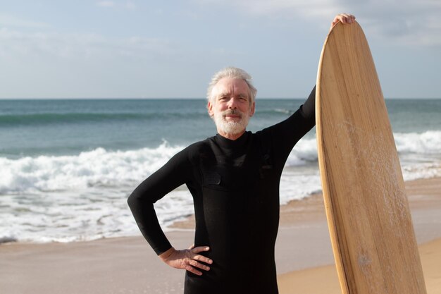 Ritratto di surfista maschio invecchiato felice che posa sulla riva del mare. Uomo dai capelli grigi in muta in piedi vicino al mare che tiene la tavola da surf con orgoglio guardando la fotocamera. Attività sportiva e riposo estivo per il concetto di persone anziane