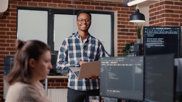 Ritratto di sviluppatore di software afroamericano che tiene il laptop guardando in alto e sorride alla telecamera. Programmatore che digita sul computer portatile accanto all'agenzia di sicurezza informatica di codifica del collega.