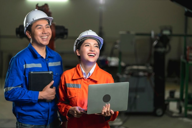 Ritratto di tecnico maschio femmina ingegnere asiatico in uniforme di sicurezza in piedi e girarsi per guardare la fotocamera e ridere sorridere con allegro e sicuro di sé sullo sfondo del posto di lavoro della fabbrica di macchinari