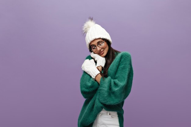 Ritratto di timida giovane donna bruna in bianco cappello lavorato a maglia e guanti Bella ragazza dai capelli lunghi in occhiali e maglione verde pone su sfondo viola