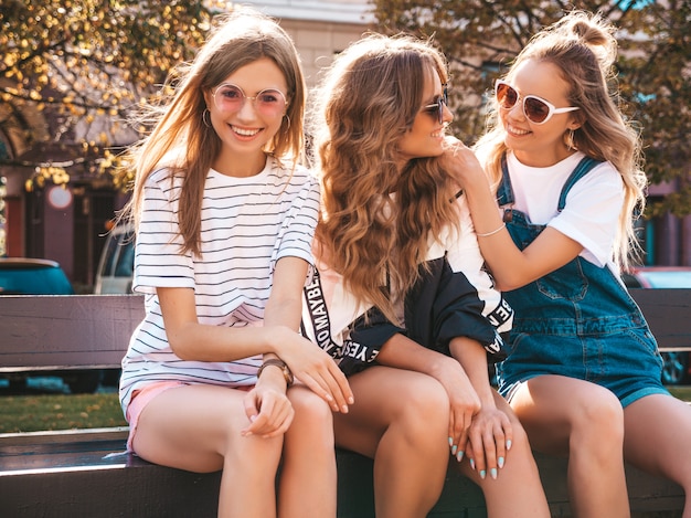 Ritratto di tre giovani belle ragazze sorridenti hipster in abiti estivi alla moda. Donne spensierate sexy seduto sulla panchina in strada. Modelli positivi divertirsi in occhiali da sole