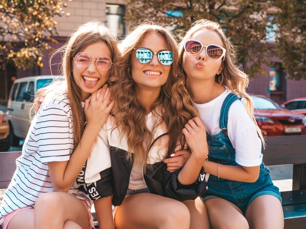 Ritratto di tre giovani belle ragazze sorridenti hipster in abiti estivi alla moda. Donne spensierate sexy seduto sulla panchina in strada. Modelli positivi divertirsi in occhiali da sole