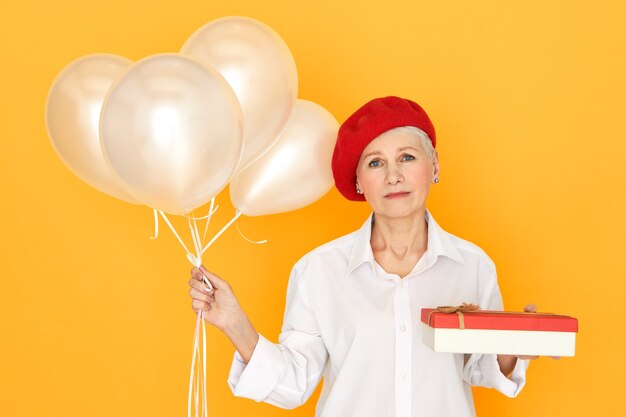 Ritratto di triste infelice donna matura in abiti eleganti in posa agaisnt sfondo giallo con scatola di cioccolata ed elio palloncini, dando regalo di compleanno, avendo sconvolto espressione depressa