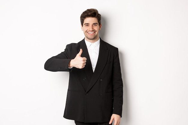 Ritratto di un agente immobiliare maschio bello e fiducioso, che mostra il pollice in su e sorride, garantisce la qualità e raccomanda la compagnia, in piedi su sfondo bianco