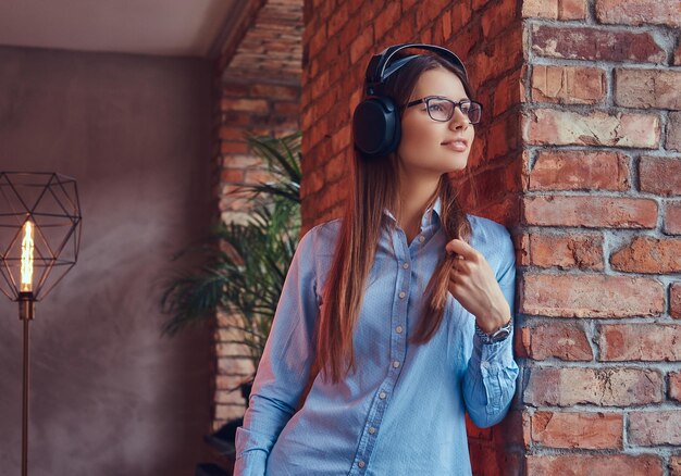 Ritratto di un'attraente bruna affascinante in occhiali e camicia blu che ascolta musica in cuffia appoggiata a un muro di mattoni in una stanza con design loft.