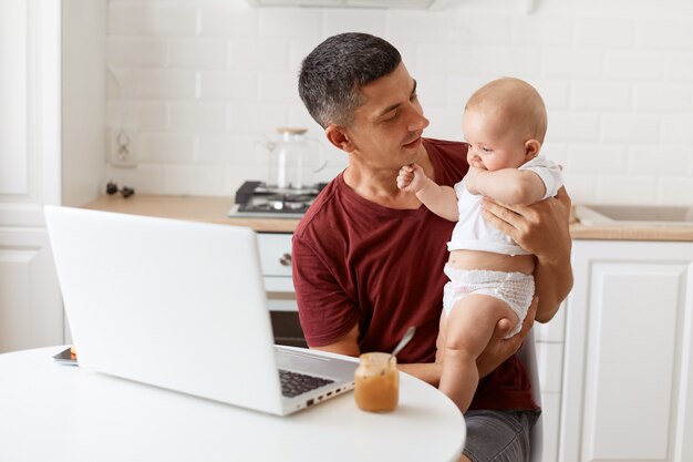 Ritratto di un attraente freelance uomo castana che indossa una maglietta marrone rossiccio in stile casual, lavora e si prende cura della sua piccola figlia, guardando il bambino con amore.