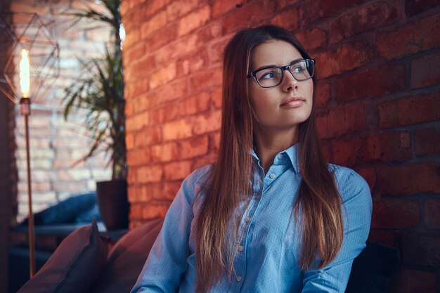 Ritratto di un'attraente ragazza bruna affascinante con gli occhiali e camicia elegante seduta sul divano al chiuso contro un muro di mattoni.