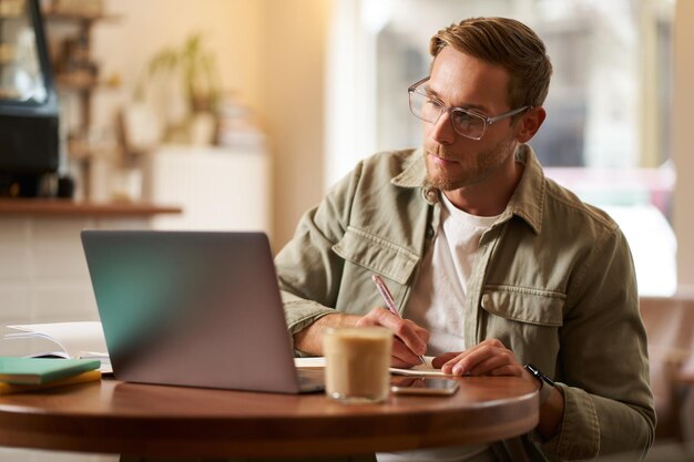Ritratto di un bel giovane con gli occhiali che studia prendendo appunti guardando lo schermo del portatile durante