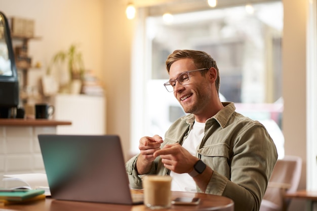 Ritratto di un bel giovane sorridente tutor online uomo d'affari che lavora in un caffè a distanza agitando la mano