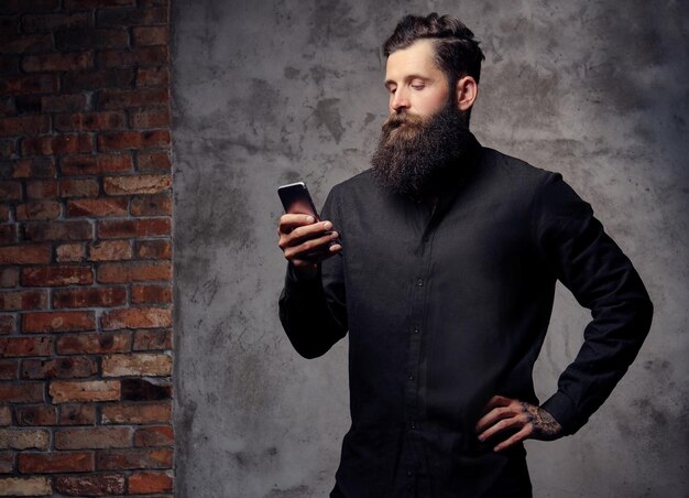 Ritratto di un bel hipster barbuto vestito con una camicia nera, utilizzando uno smartphone, in piedi in uno studio. Isolato su uno sfondo scuro.