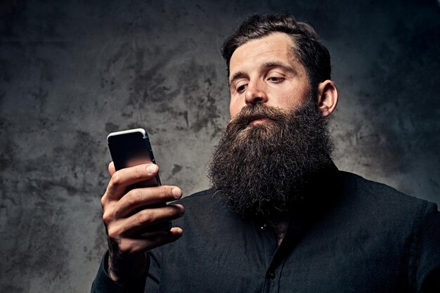 Ritratto di un bel hipster barbuto vestito con una camicia nera, utilizzando uno smartphone, in piedi in uno studio. Isolato su uno sfondo scuro.