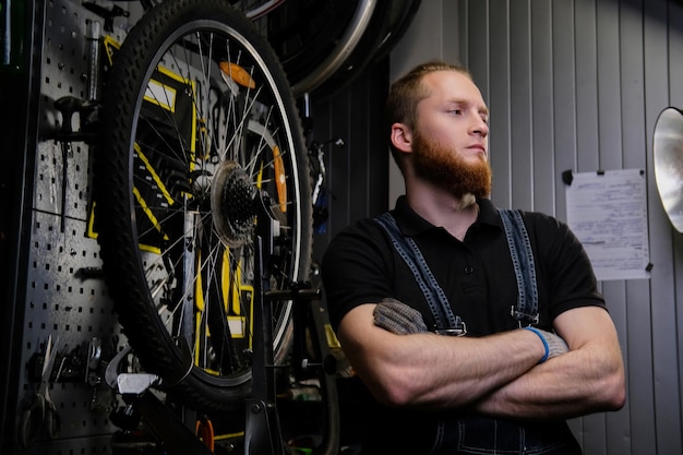 Ritratto di un bel maschio rosso con barba e taglio di capelli che indossa una tuta di jeans, in piedi vicino alla ruota di bicicletta in un'officina.