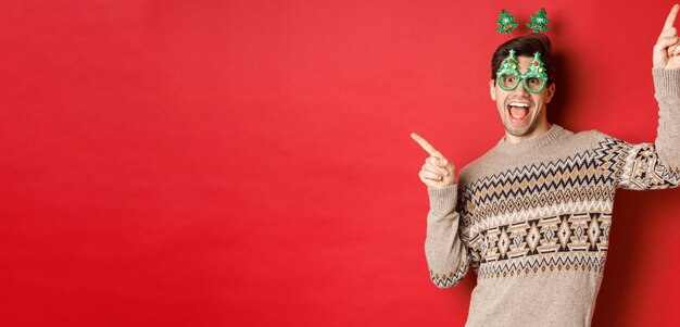 Ritratto di un bel ragazzo gioioso con gli occhiali da festa e un maglione natalizio che balla e punta il dito...