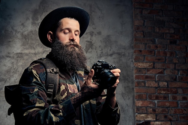 Ritratto di un bel viaggiatore barbuto con cappello e giacca mimetica, con uno zaino e braccia tatuate, tiene una macchina fotografica. Isolato su uno sfondo scuro.