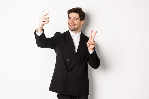 Ritratto di un bell'uomo che si fa selfie alla festa di capodanno, indossa un abito, scatta foto sullo smartphone e mostra il segno della pace, in piedi su sfondo bianco