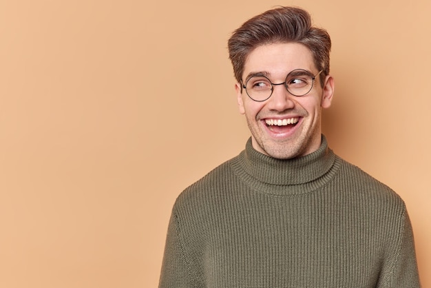 Ritratto di un bell'uomo con un ampio sorriso sincero distoglie lo sguardo si sente bene vede qualcosa di divertente indossa occhiali rotondi maglione casual isolato su sfondo beige con copia spazio per la tua pubblicità