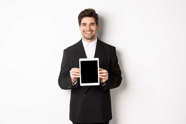 Ritratto di un bell'uomo d'affari in abito alla moda, che mostra lo schermo del tablet digitale e sorride, in piedi su sfondo bianco