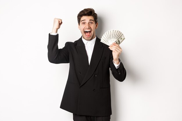 Ritratto di un bell'uomo d'affari in abito nero, che vince denaro e si rallegra, alzando la mano con eccitazione, in piedi su sfondo bianco.