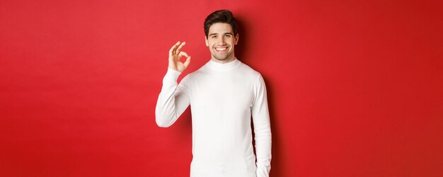 Ritratto di un bell'uomo felice e soddisfatto, che indossa un maglione bianco, mostra un segno giusto e sorride, approva qualcosa di buono, consiglia un negozio, in piedi su sfondo rosso.