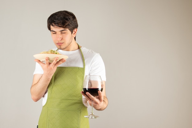 Ritratto di un bell'uomo in grembiule che tiene in mano un piatto con noodles e un bicchiere di vino.