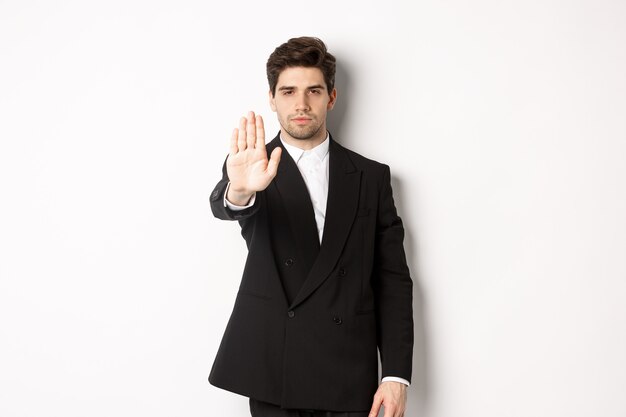 Ritratto di un bell'uomo serio in abito formale, che allunga la mano per fermarti, proibire l'azione, vietare e non essere d'accordo con qualcosa, in piedi su sfondo bianco