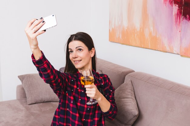 Ritratto di un bicchiere di vino sorridente della tenuta della giovane donna che prende selfie sul telefono cellulare a casa