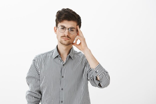 Ritratto di un collega maschio serio concentrato in occhiali rotondi, guardando in basso e tenendo la tempia con il dito indice, concentrandosi mentre pensa, preparando un piano su come evitare situazioni scomode