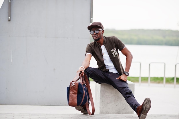 Ritratto di un elegante uomo afroamericano seduto su occhiali da sole e berretto all'aperto Uomo nero di moda di strada