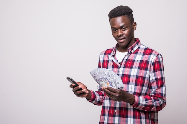 Ritratto di un giovane africano soddisfatto vestito con una camicia a quadri che tiene il telefono cellulare e un mazzo di banconote di denaro isolato
