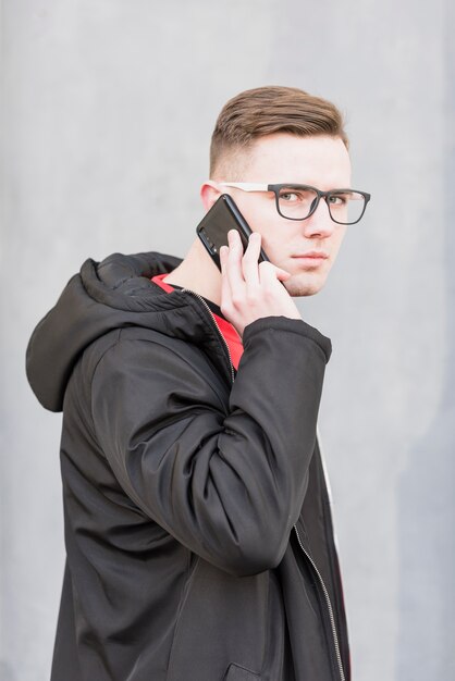 Ritratto di un giovane attraente con gli occhiali che parla sul telefono cellulare contro il contesto grigio