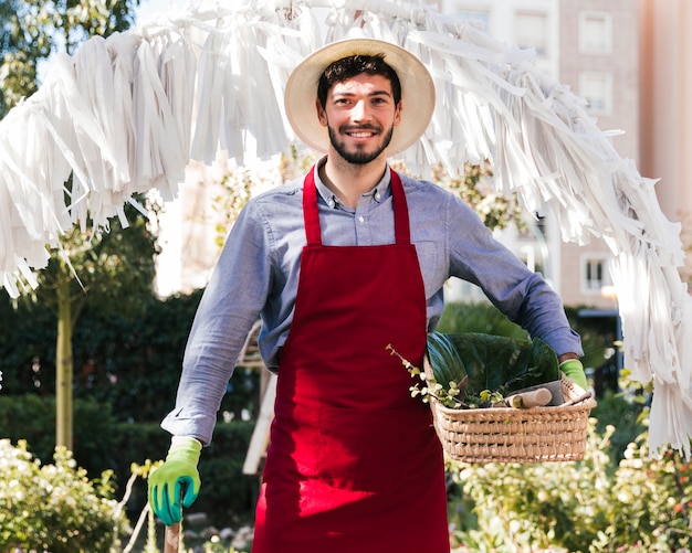 Ritratto di un giovane giardiniere maschio sorridente che tiene strumento e canestro di giardinaggio che esaminano macchina fotografica