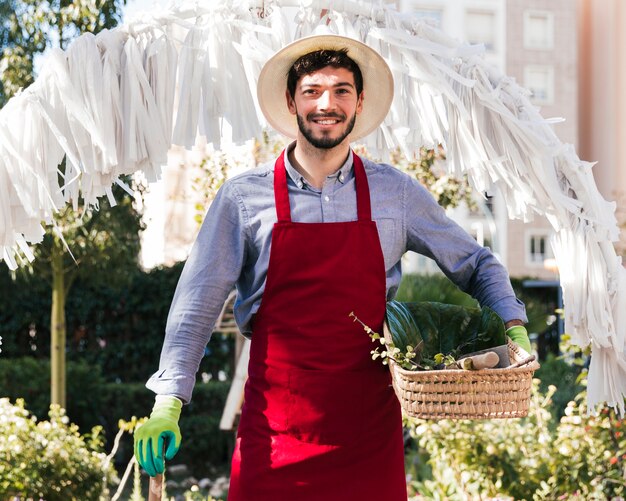 Ritratto di un giovane giardiniere maschio sorridente che tiene strumento e canestro di giardinaggio che esaminano macchina fotografica