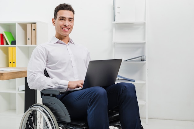 Ritratto di un giovane sorridente che si siede sulla sedia a rotelle con il computer portatile che guarda l&#39;obbiettivo