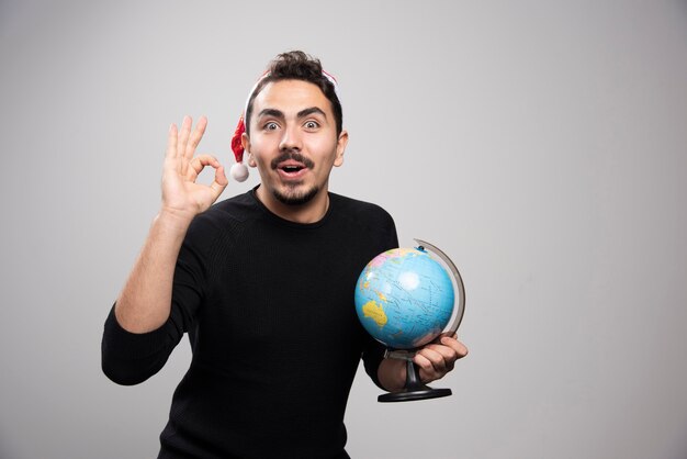 Ritratto di un giovane uomo con il cappello di Babbo Natale che mostra il segno giusto e che tiene un globo.