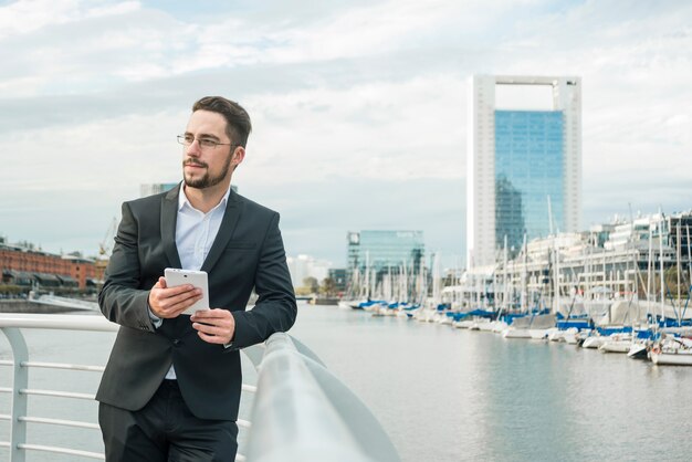 Ritratto di un giovane uomo d&#39;affari in piedi vicino al porto tenendo in mano il telefono cellulare in cerca di distanza