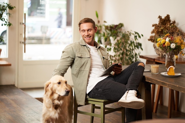 Ritratto di un giovane uomo felice e sorridente che visita un caffè seduto in un caffè con il suo cane che accarezza l'oro