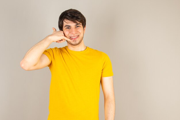 Ritratto di un giovane uomo in piedi e facendo segno di telefonata contro il grigio.