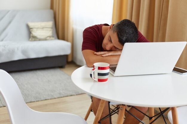 Ritratto di un libero professionista stanco che si addormenta sul tavolo vicino al computer portatile dopo aver lavorato online, indossando una maglietta marrone rossiccio in stile casual, dormendo, appoggiandosi alle mani, posando in un luminoso soggiorno vicino alla finestra.