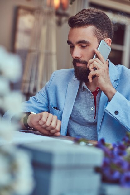 Ritratto di un maschio barbuto alla moda con un taglio di capelli alla moda, parlando al telefono, guardando un orologio da polso, seduto in un caffè all'aperto.