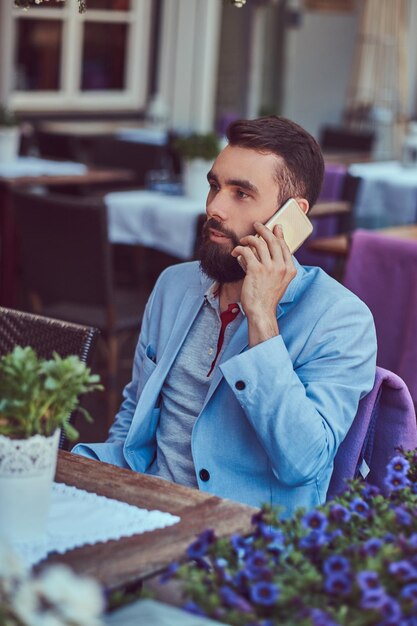 Ritratto di un maschio barbuto alla moda con un taglio di capelli alla moda, parlando al telefono, seduto in un caffè all'aperto.