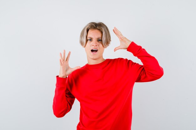 Ritratto di un maschio biondo adolescente che allunga le mani in modo aggressivo in un maglione rosso e sembra una vista frontale furiosa
