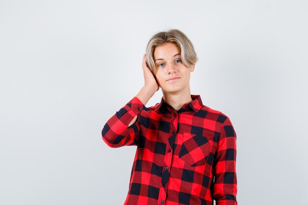 Ritratto di un maschio biondo adolescente che copre l'orecchio con la mano in una camicia casual e guarda una vista frontale pensierosa