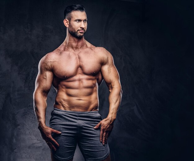 Ritratto di un maschio enorme alto senza maglietta con un corpo muscoloso con un taglio di capelli alla moda e la barba, in pantaloncini sportivi, in posa in uno studio. Isolato su uno sfondo scuro.