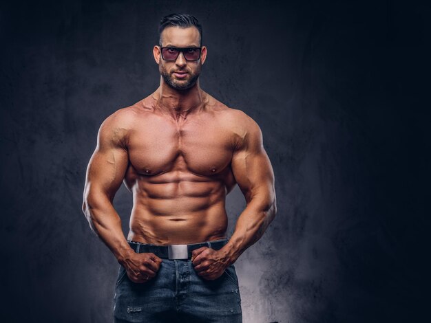 Ritratto di un maschio enorme alto senza maglietta con un corpo muscoloso con un taglio di capelli e barba alla moda, in occhiali da sole e jeans, in posa in uno studio. Isolato su uno sfondo scuro.