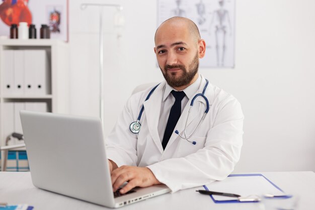 Ritratto di un medico specialista che guarda nella telecamera che lavora nella sala conferenze analizzando le competenze in materia di malattia utilizzando il computer portatile. Medico che prescrive il trattamento sanitario dei farmaci delle pillole