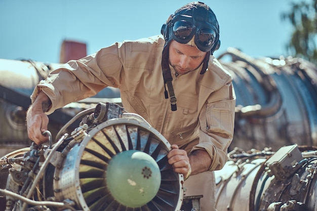 Ritratto di un pilota-meccanico in uniforme e casco volante, che ripara la turbina dell'aeroplano smontata in un museo a cielo aperto.