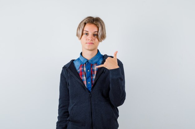 Ritratto di un ragazzo adolescente carino che mostra il gesto del telefono in camicia, felpa con cappuccio e sembra fiducioso in vista frontale