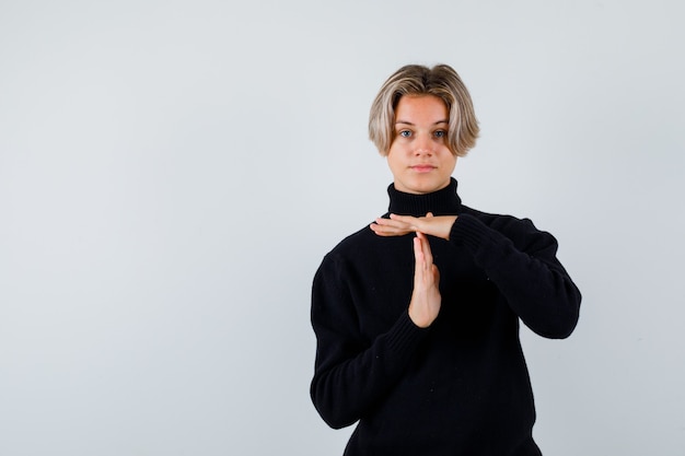 Ritratto di un ragazzo adolescente carino che mostra il gesto della pausa temporale con un maglione a collo alto e sembra una vista frontale sicura