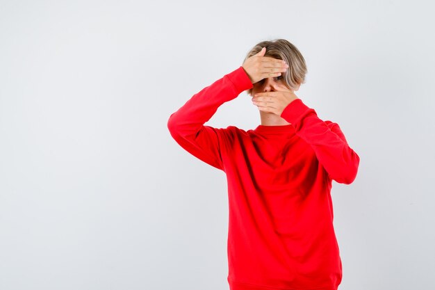 Ritratto di un ragazzo adolescente che guarda attraverso le mani in un maglione rosso e sembra spaventato vista frontale